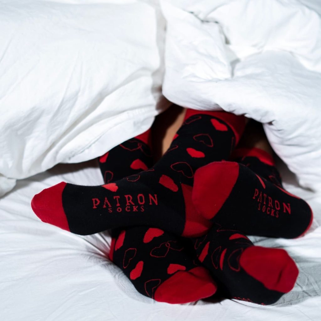 4 Beine unter Bettdecke mit roten Herz Socken_PATRON SOCKS